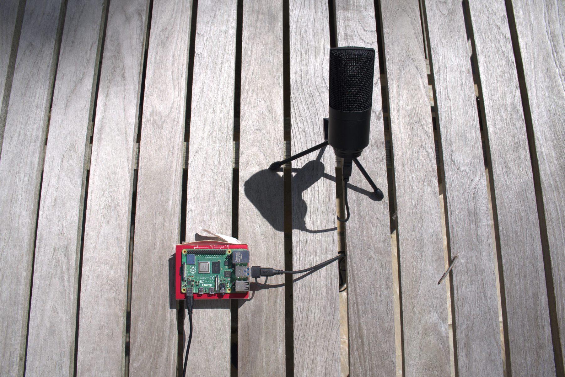 HeliTracker device: a Raspberry Pi in a red case connected to a USB microphone on a tripod, photographed from above on a wooden deck